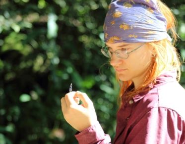 Young woman in bandana looks at a butterfly perched on her hand, against a lush background.