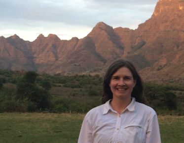 Woman with long hair, wearing a light-colored shirt, smiles at the camera, in front of a background of mountains lit up with alpenglow.