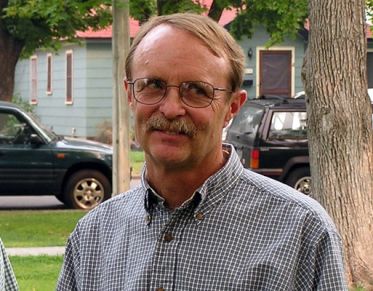 Mustachioed man, wearing glasses and a checked shirt, smiles at someone off-camera, in front of a mid-summer, treelined, residential street.