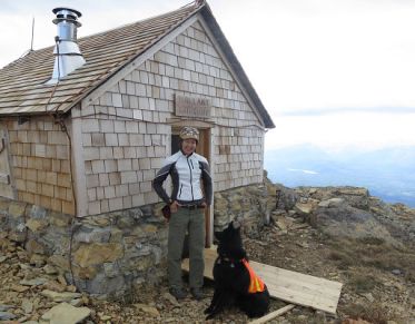 Woman with curly hair, wearing a gray and white jacket and a hat, smiles at the camera from in front of a shingled alpine hut, in rocky, mountainous terrain.