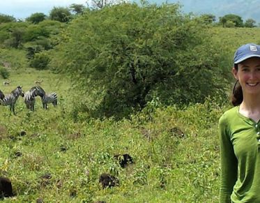 Woman wearing ball cap smiles at the camera in front of a small herd of zebra on a lush African savannah.