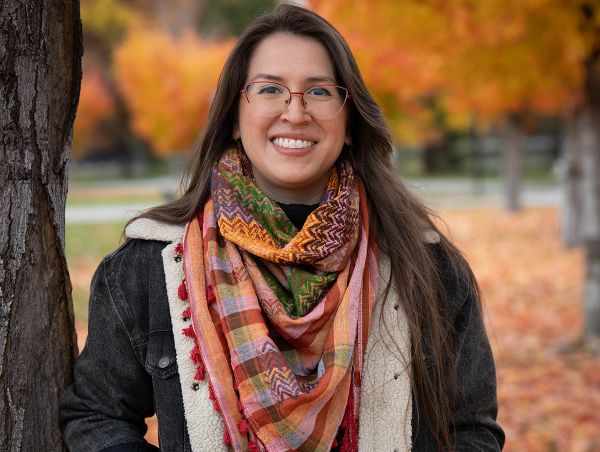 Young woman wearing jacket and colorful scarf stands amidst maple trees in full autumn color.