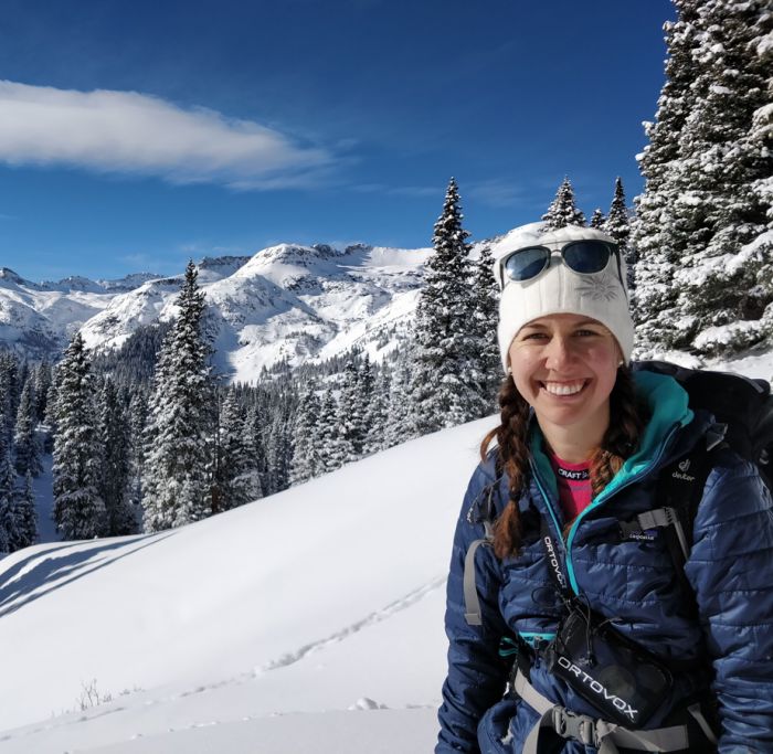 Amy Katz smiles at the camera while outside in the snow-covered mountains. She is wearing a white knit hat. A large, snow-covered peak looms in the background.