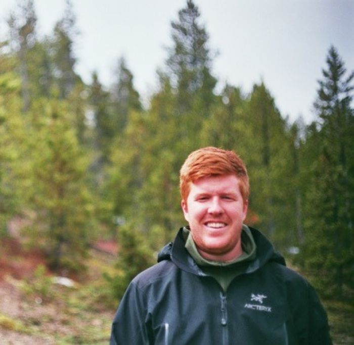 Sawyer Connelly smiles at the camera while standing in a forest. He wears a black Arcteryx jacket