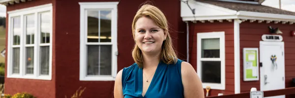 A woman stands in front of a one-room schoolhouse 
