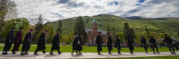 row of graduates in graduation garb walking down the middle of the oval with University Hall and the M trail in the background