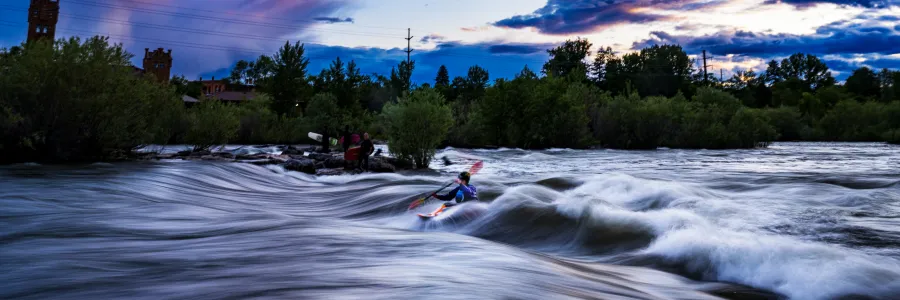 A kayaker surfs Brennan's Wave on the Clark Fork River in downtown Missoula under a summer sunset with moody clouds.