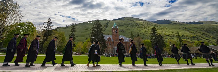 row of graduates in graduation garb walking down the middle of the oval with University Hall and the M trail in the background