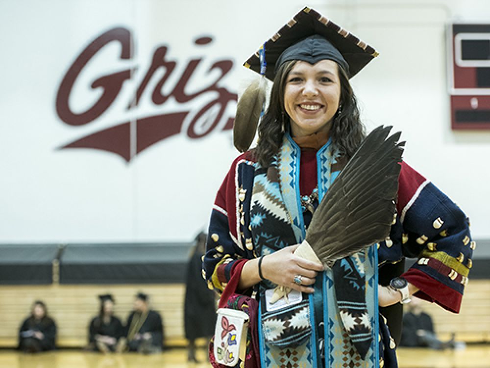 Rose Bear Don’t Walk stands before the camera in her graduation regalia in front of a wall that reads: Griz