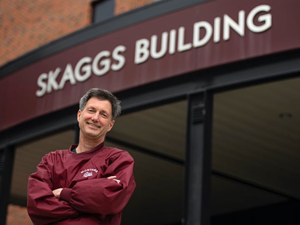 A male stands with arms folded, wearing a Griz shirt and smiling in front of Skaggs Building facade