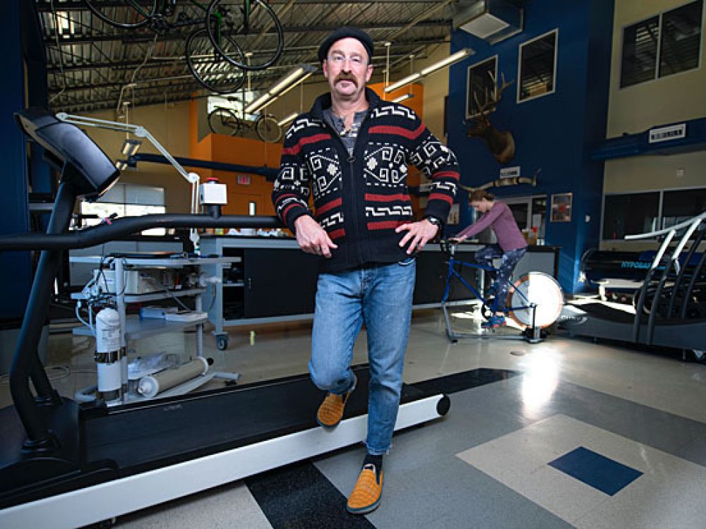 Brent Ruby stands in his UM lab with a subject pedaling in the background.