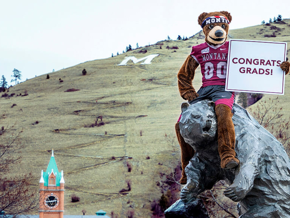 Monte the mascot sits on top of the Grizzly Bear Statue while holding a sign that reads Congrats, Grads! Mounti Sentinel in the background