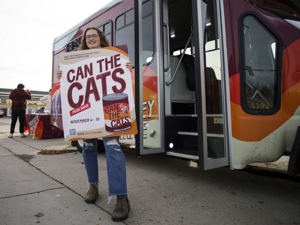 A student holds a Can the Cats sign outside a bus.