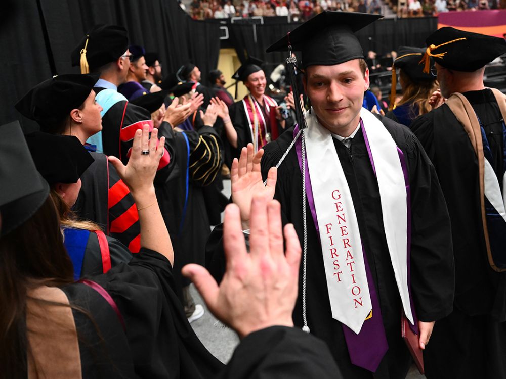 A UM student high-fives a fellow classmate at Commencement. He wears a sash that says "First-Gen."