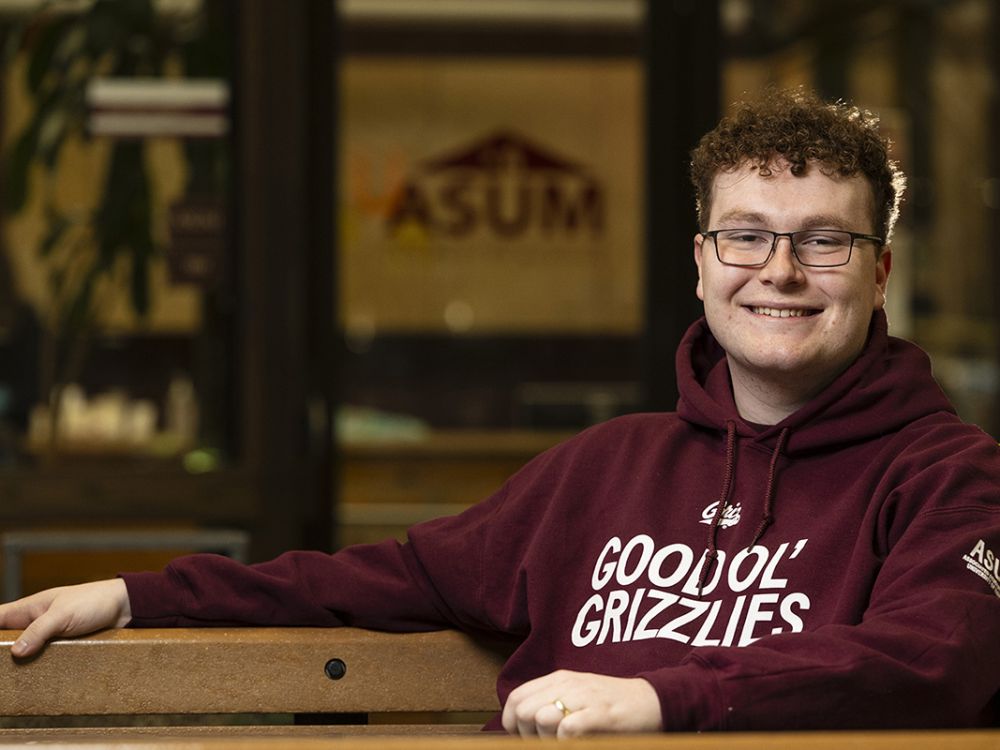 Liam Edwards sits in the University Center wearing a "Good Ol' Grizzlies" sweatshirt. The ASUM logo can be seen on a window in the background.