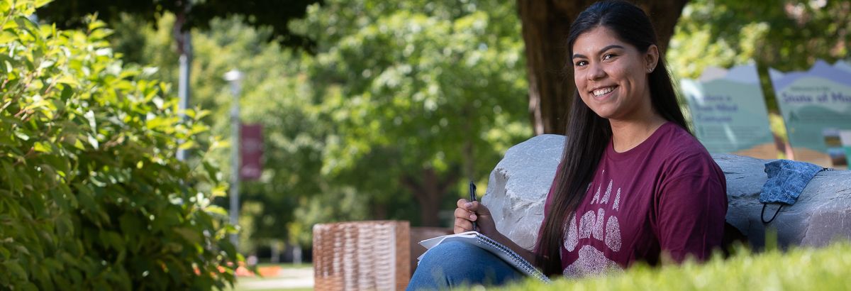 smiling student outside sitting against a rock with a notebook and pen