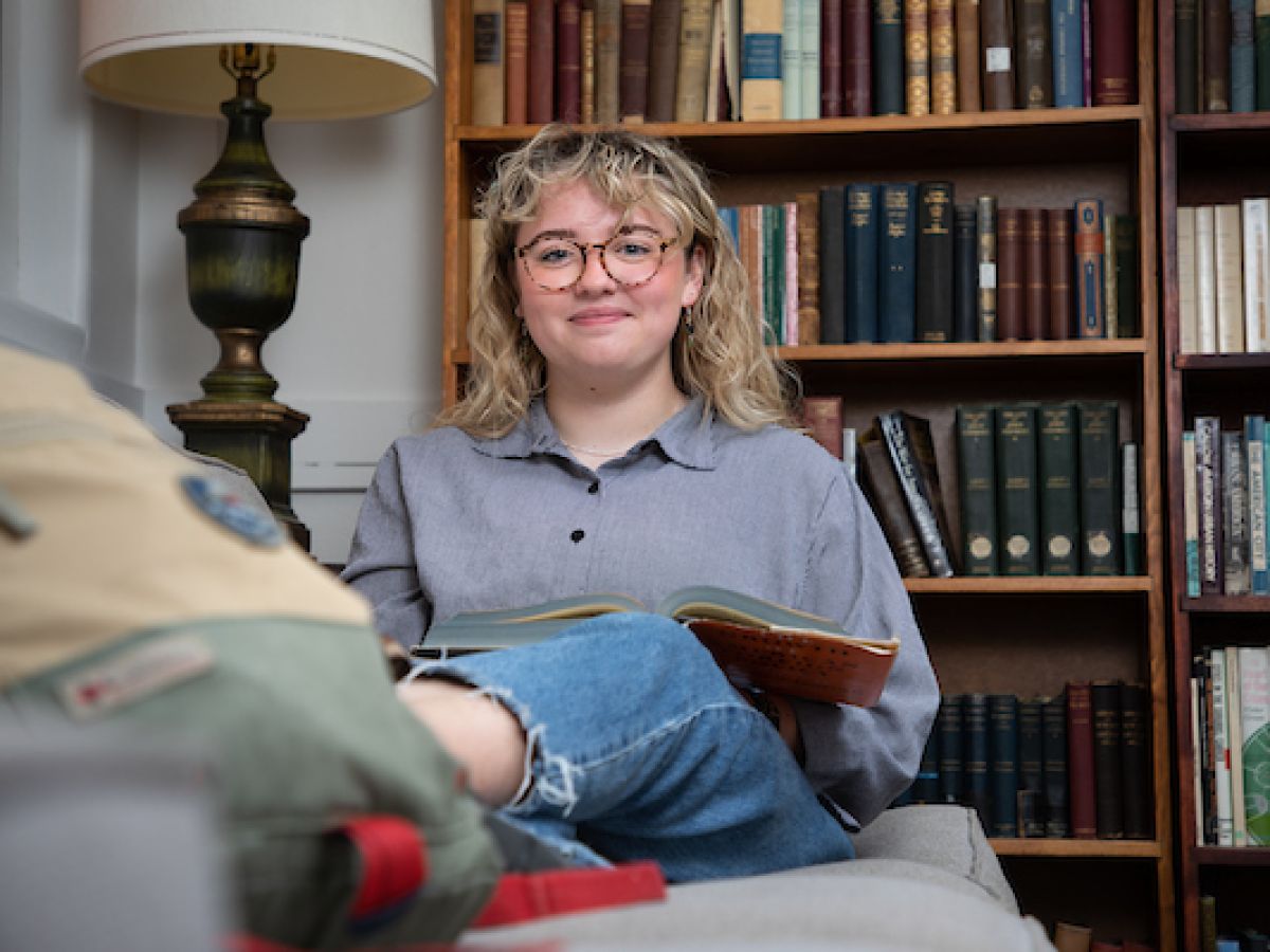 Maddie sits on a couch with a book and smiles at the camera