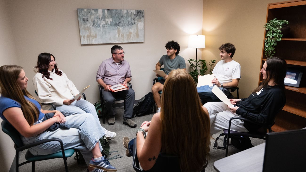 A group of college students sits in a circle in a bright room, engaging in a guided group therapy session. Some students are speaking while others listen attentively, creating a supportive and inclusive environment.