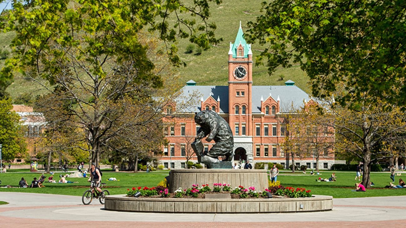 The University of Montana Oval on a sunny day, with students relaxing on the grass and the bronze Grizzly Bear statue in the foreground, and Main Hall with its clock tower in the background.