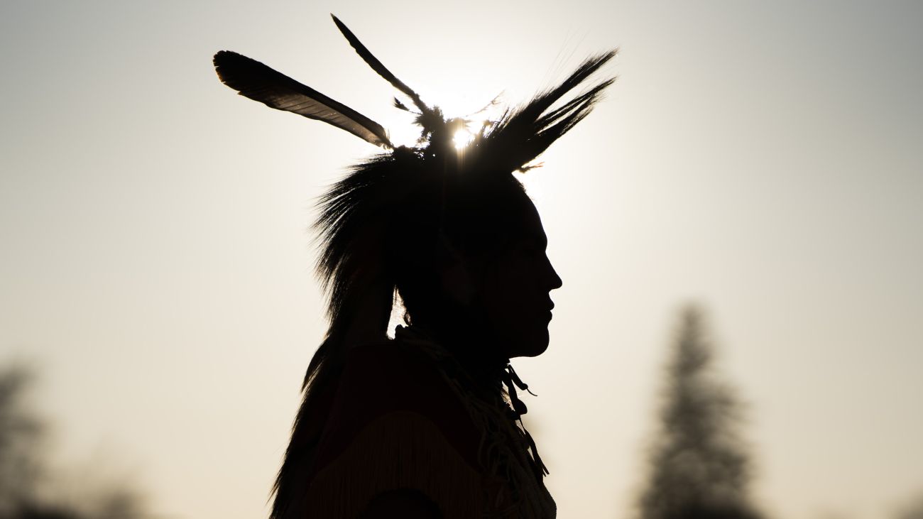 Silhouette of a dancer in regalia showing a head roach and feathers with the sun glistening behind