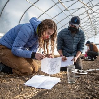 students kneel on dirt in greenhouse and look at pages of homework