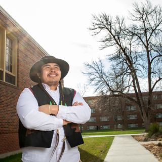 Zachariah Rides at the Door stands outside a brick building on campus. He wears a black hat and vest and holds his arms across his chest as he smiles.