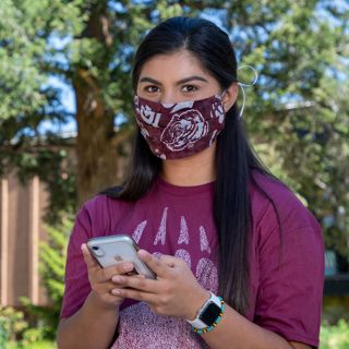 Female student wearing a cloth face covering holds her phone while standing in front of the UC