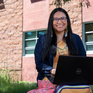 A student smiles at the camera while sitting outside the Payne Family Native American Center. There is a laptop on her lap.