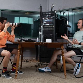 Two male students in audio booth, one taking a photo of the other talking into a microphone.