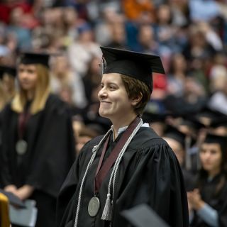 A graduate wears a cap and gown jumps in the air in front of Main Hall
