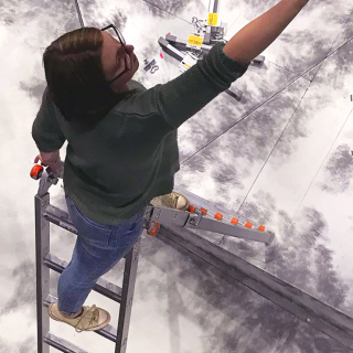 A design student paints a set atop a ladder, shot from above.