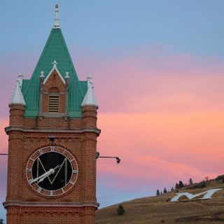 The clock tower of Main Hall at UM with Mount Sentinel in the background and a pink cloud in the sky