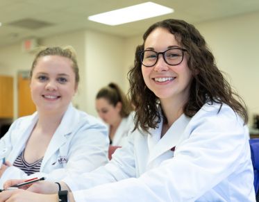 Two pharmacy students in lab coats smiling from their classroom seats