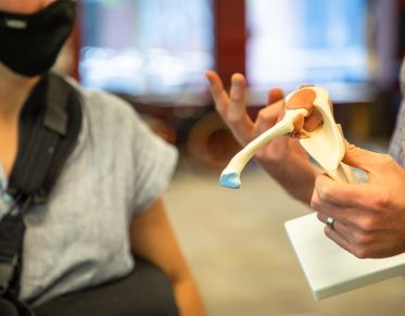 Faculty teaching with model of bone and muscle as student wearing a shoulder brace observes