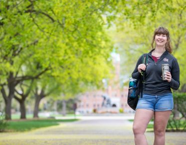 Female student holds backpack and Montana coffee mug in front of trees, the griz statue, and Main Hall