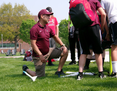 A role model kneels in grass as they demonstrate a rocket-launching activity for a group of middle school students.