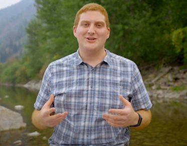 UM student Jonathan Karlen looks at the camera while speaking as he sits along the banks of the Clark Fork River that flows past campus.