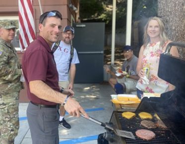 Military and Veterans Services Director Patrick Barkey smiles at the camera while grilling burgers. Four students hang out in the background.