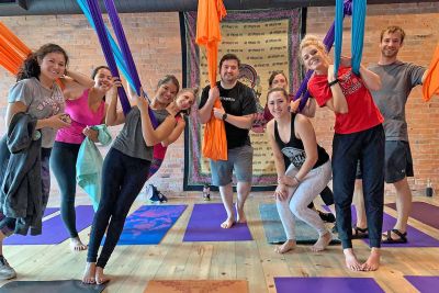 There are nine people in a yoga studio with colorful aerial yoga bands extending from the ceiling and yoga mats on the floor and a tapestry in the background 