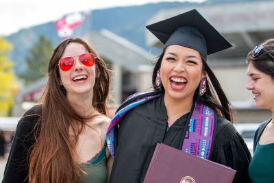 Three women smile, with one in graduation attire. 