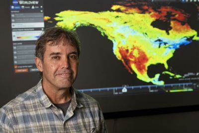 A picture of John Kimball standing before a multicolored video map of the Earth.