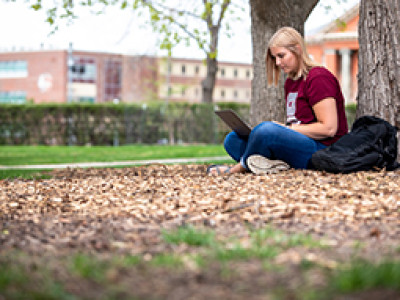 A student sits on the ground beneath a tree, working on a laptop in a park-like setting