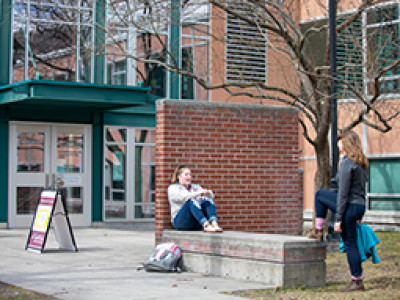 Two students are engaging in conversation outside a modern building on a university campus, with one sitting on a low wall and the other standing nearby