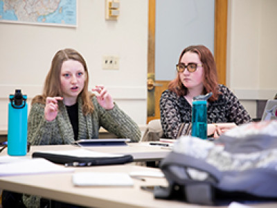 Two students engaged in a discussion during a classroom setting, one gesturing while speaking and the other attentively listening