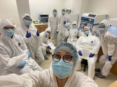 People in white full-body biohazard suits and blue face masks look at camera in a lab 