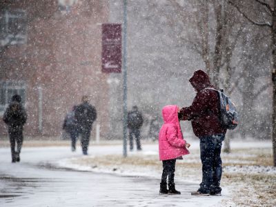 A caregiver stops to help a child fix their bright pink coat in the snow