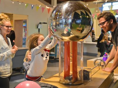 A child reaches for a large, silver ball that is part of a science exhibit. An educator leans forward, engaging with them.