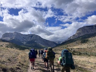 students carrying backpacks hike away from camera toward mountains
