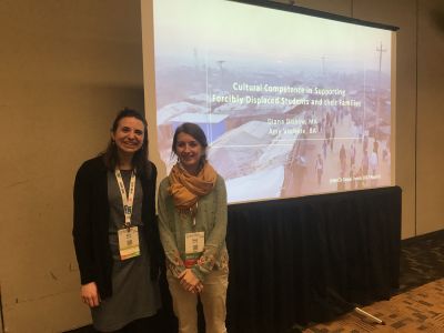 Two Graduate students standing in front of a PowerPoint entitled "Cultural Competence in Supporting Forcibly Displaced Students and Their Families""