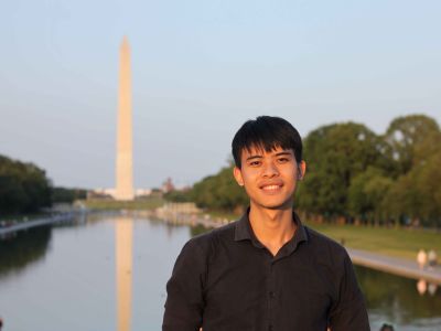 A young man smiling in front of the Washington Monument, reflecting in the nearby water at sunset